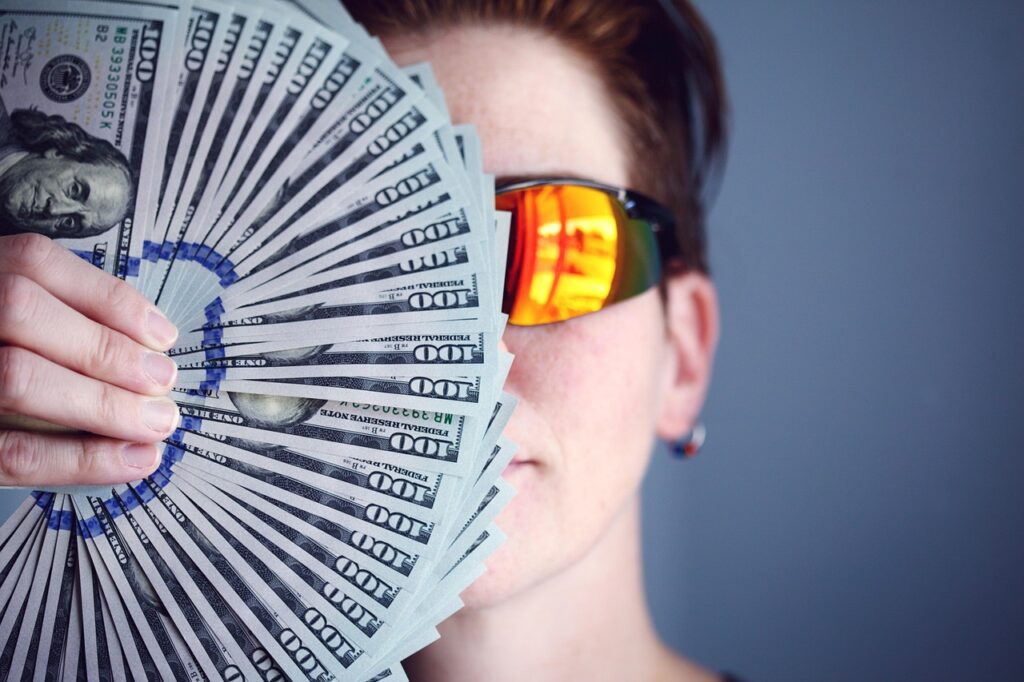 A person wearing orange sunglasses holding a fan of one hundred dollar bills.