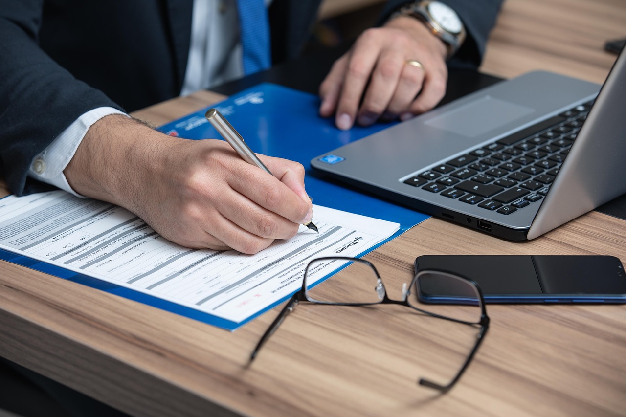 Close-up of a businessman signing papers on a desk next to a laptop and glasses.